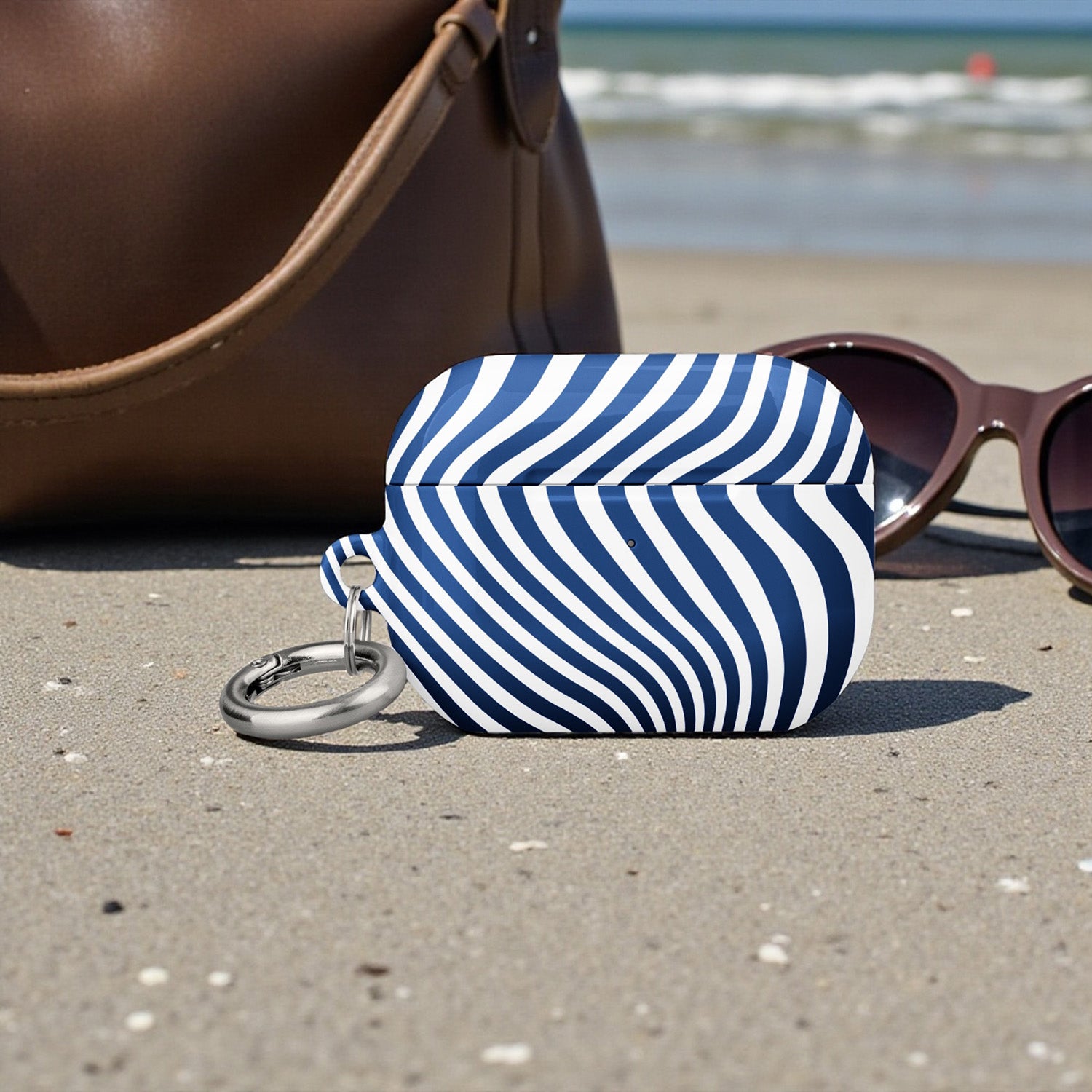 Blue and white striped pouch with keychain on a sandy beach with sunglasses and handbag in the background.